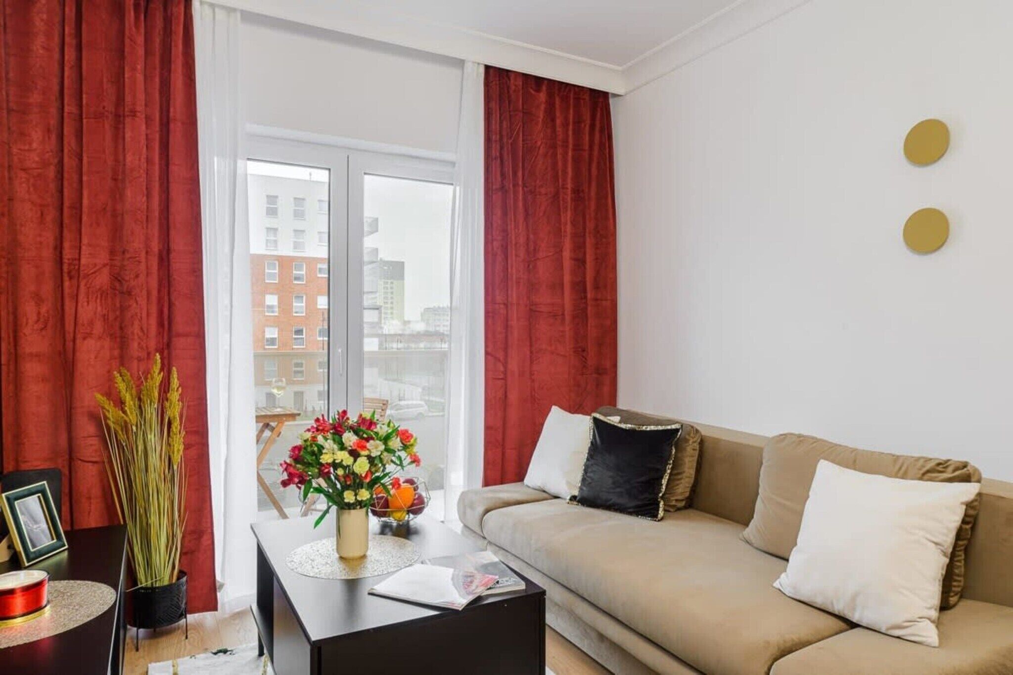 Ein Wohnzimmer mit einem beigefarbenen Sofa, roten Vorhängen, einer Vase mit Blumen und Blick auf Gebäude durch ein großes Fenster.