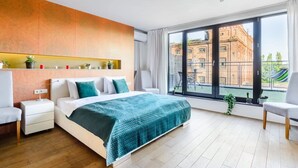 Un dormitorio moderno con una cama grande, una mesita de noche, una silla y vista a un edificio a través de un ventanal amplio.