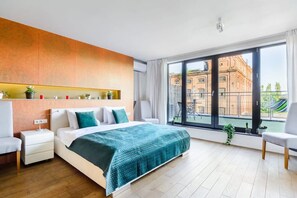 Ein modernes Schlafzimmer mit einem großen Bett, einem Nachttisch, einem Sessel und Blick auf ein Gebäude durch ein großes Fenster.