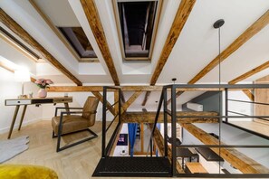 Ein Loft-Zimmer mit freiliegenden Holzbalken, einem Schreibtisch, einem Sessel und einem kleinen Bücherregal.
