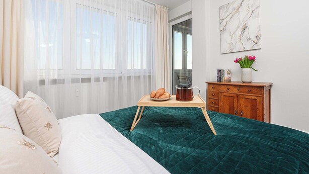 Een slaapkamer met een bed, een nachtkastje met een schaal croissants en een theepot, een houten dressoir en een raam met dunne gordijnen.