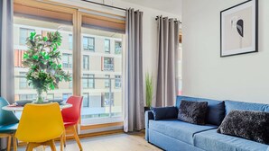 Ein modernes Wohnzimmer mit einem blauen Sofa, einem Esstisch mit Stühlen und einem großen Fenster mit Vorhängen.