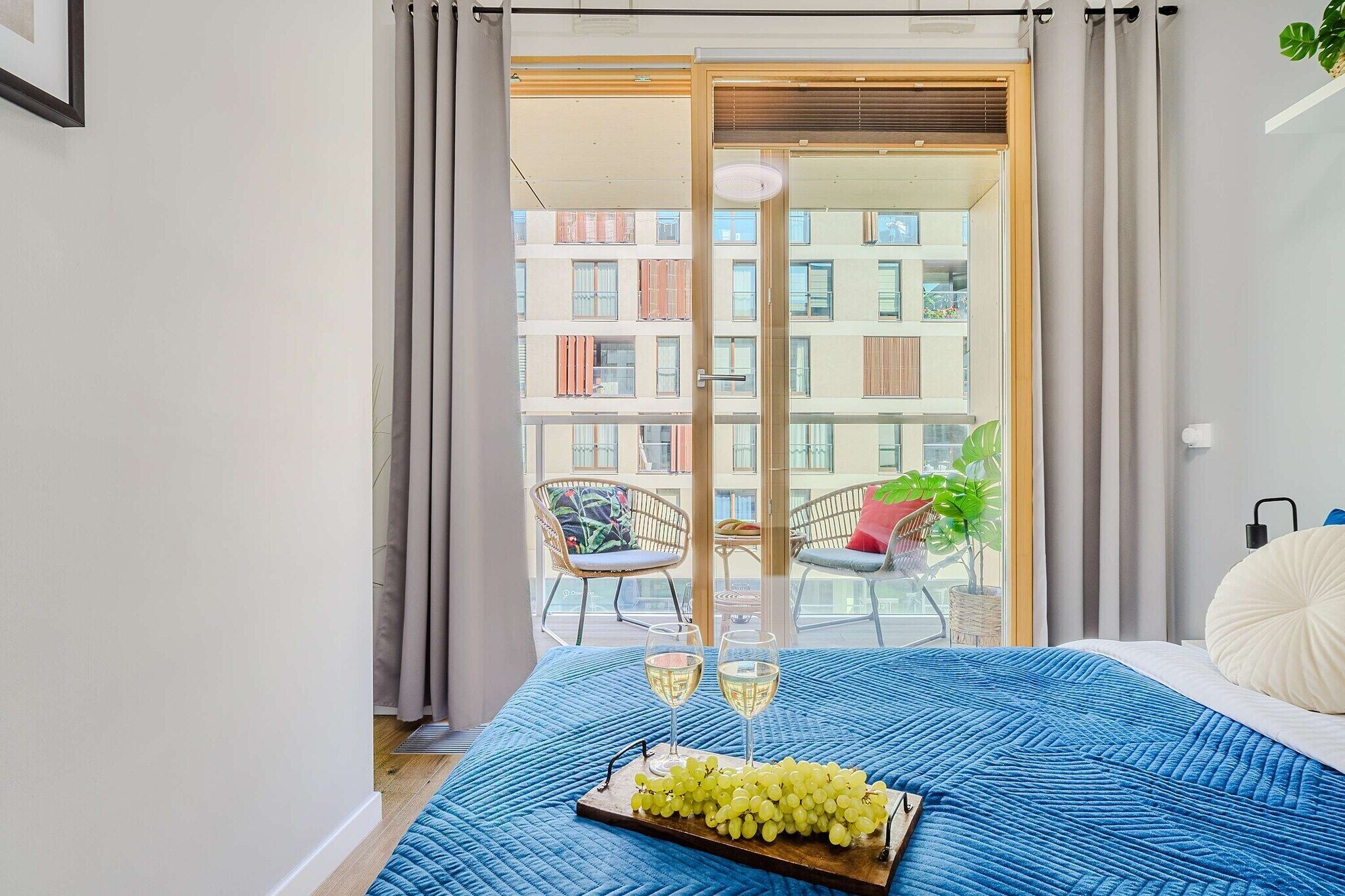Ein Schlafzimmer mit einem Bett, einem Glastisch mit zwei Weingläsern und einer Traubentafel, sowie ein Balkon mit Stühlen und einer Pflanze.
