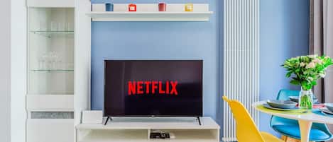 Una sala moderna con una televisión de pantalla plana mostrando 'NETFLIX', un mueble blanco para la televisión, una mesa de comedor con sillas coloridas y un radiador.