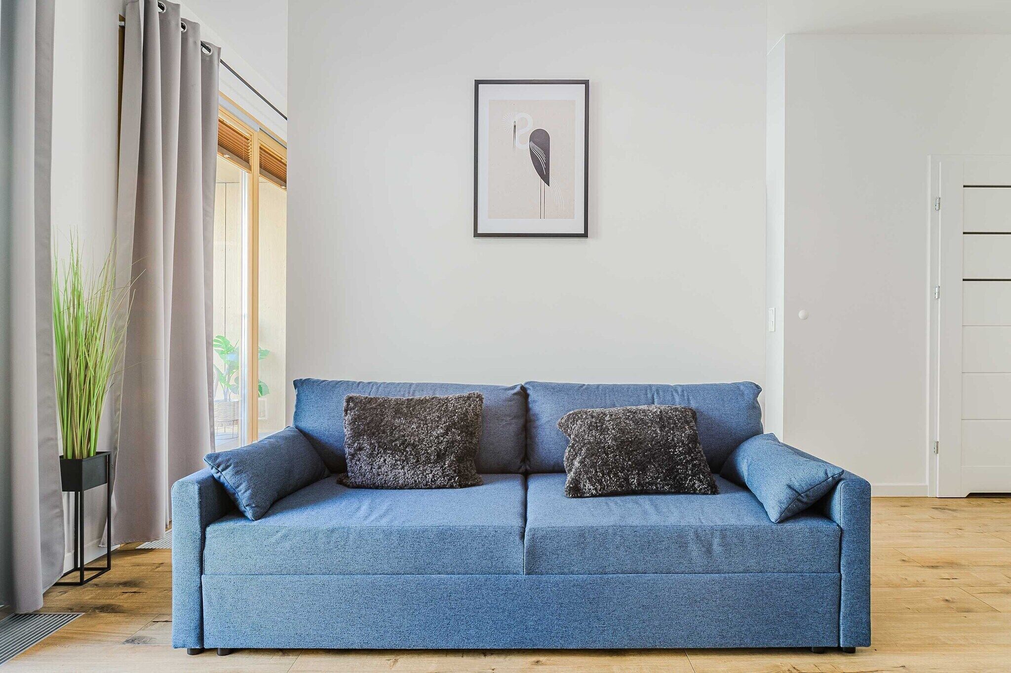 A blue sofa with two textured pillows, a framed artwork on the wall, and a potted plant beside a window.