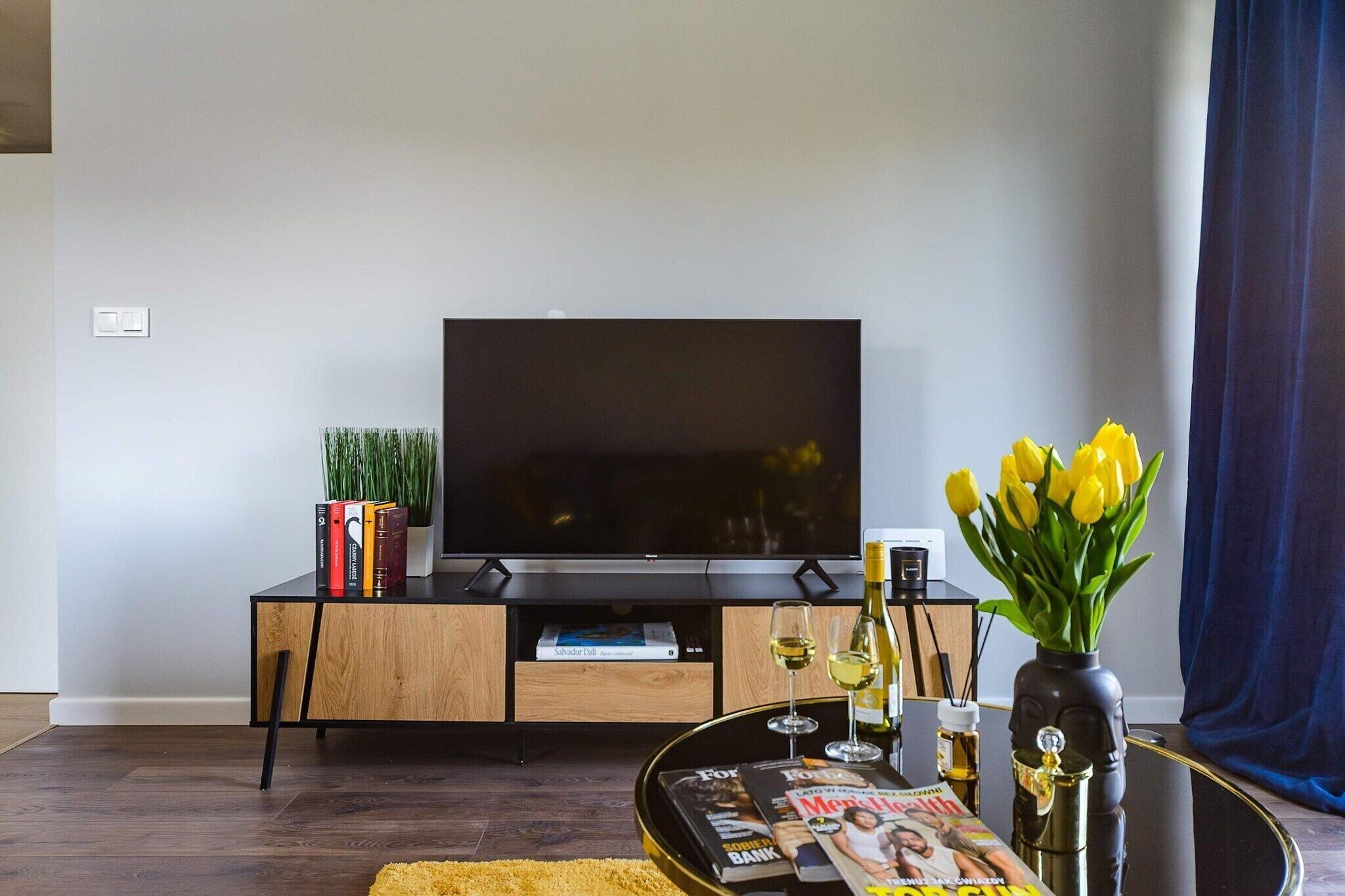 A modern living room with a flat-screen TV, wooden entertainment unit, books, a magazine, a vase with yellow flowers, and a glass of wine.