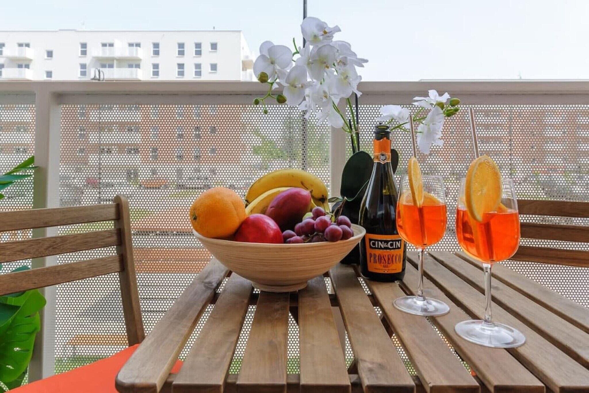 A wooden table with a bowl of fruit, a bottle of Prosecco, and two glasses of orange wine on a balcony.