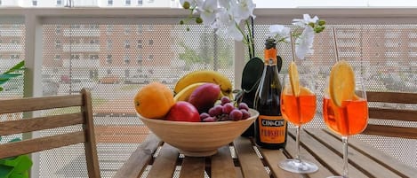 Una mesa de madera con un cuenco de fruta, una botella de Prosecco y dos copas de vino naranja en un balcón.