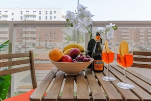Une table en bois sur laquelle se trouvent un saladier de fruits, une bouteille de Prosecco et deux verres de vin orange, le tout posé sur un balcon.