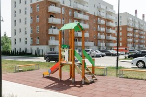 A playground with a slide and climbing structure, surrounded by a parking lot and a multi-story residential building. - 3-Bedroom Łódź Apartment with Balcony (Łódź)
