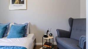 Ein Schlafzimmer mit einem Bett, einem grauen Sessel, einem kleinen Tisch mit Tasse und Büchern sowie einem gerahmten Bild an der Wand.