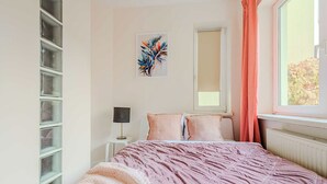 Et soveværelse med en seng, et natbord, en lampe, et vindue med gardiner og en vægmonteret hylde.