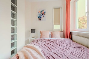Ein Schlafzimmer mit einem Bett, einem Nachttisch, einer Lampe, einem Fenster mit Vorhängen und einem an der Wand befestigten Regal.