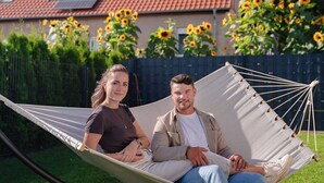 Een hangmat met twee personen, een huis met rode dakpannen en zonnebloemen op de achtergrond.