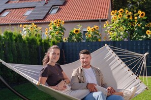 Eine Hängematte mit zwei Personen, ein Haus mit rotem Ziegeldach und Sonnenblumen im Hintergrund.