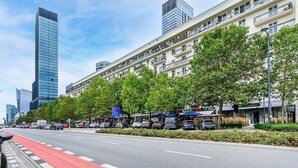 Eine Straße mit Bäumen und parkenden Autos, im Hintergrund sind Gebäude zu sehen.