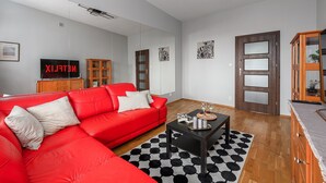Ein modernes Wohnzimmer mit einem roten Ecksofa, einem schwarz-weißen Teppich, einem Couchtisch mit Dekoration, einer Holzkommode und einem Fernseher, der das Wort 'NETFLIX' anzeigt.