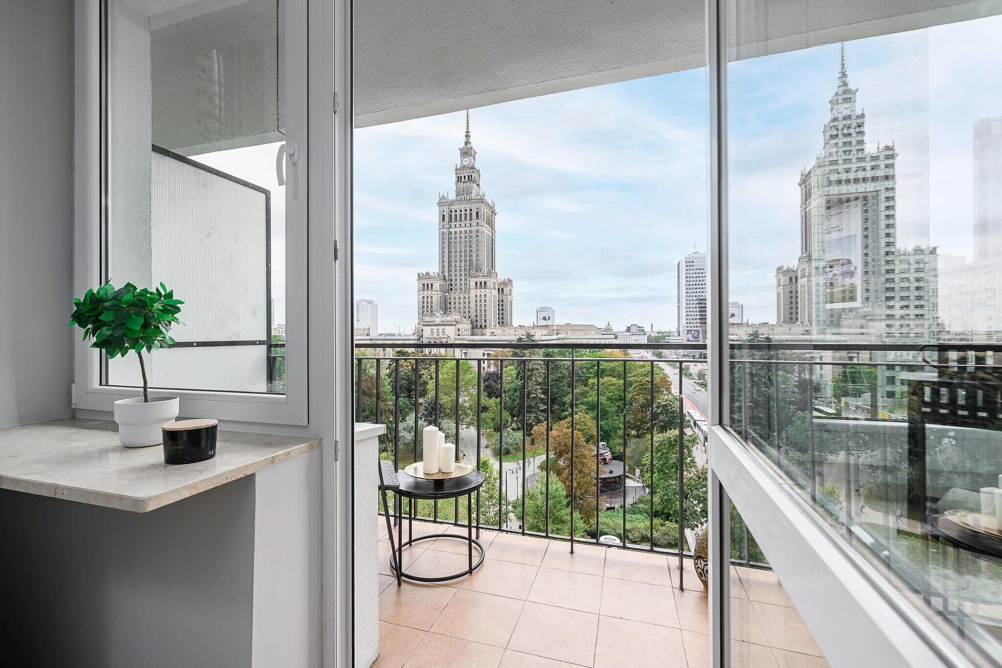 Een balkon met uitzicht op stadsgebouwen, een bloempot en een kleine tafel met kaarsen.