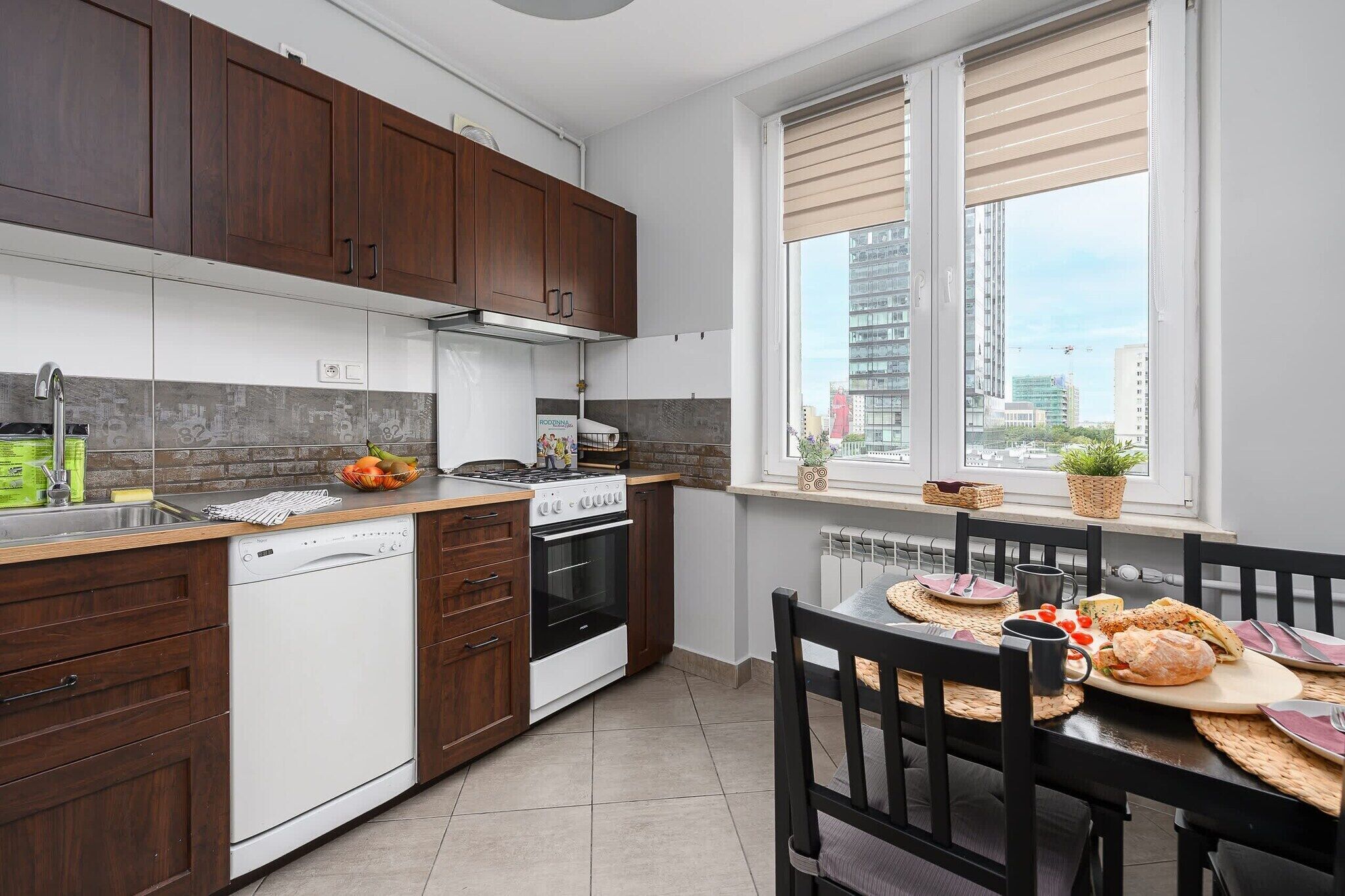 A modern kitchen with dark wooden cabinets, a white dishwasher, and a stove. There is a dining table with a black chair and a window with blinds.