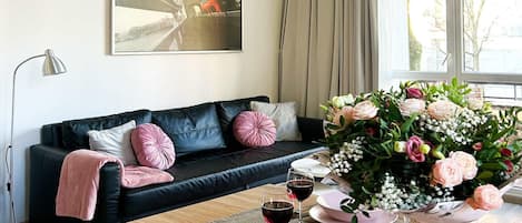 Un salon moderne avec un canapé en cuir noir, des coussins roses, une table à manger avec de l’assiette rose et des verres à vin, et un grand arrangement floral.
