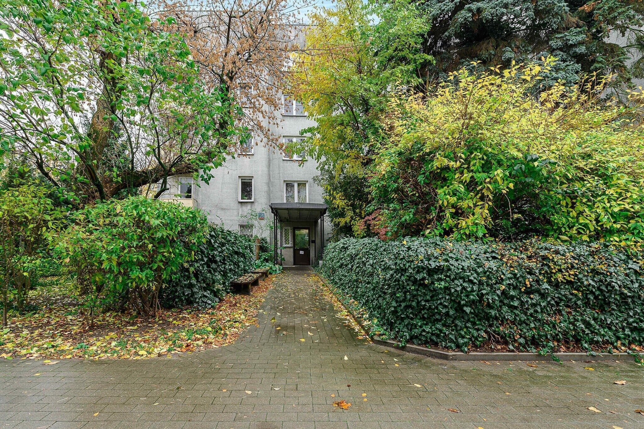 Un chemin menant à un bâtiment doté d’une entrée abritée, entouré d’un feuillage dense et d’arbres.