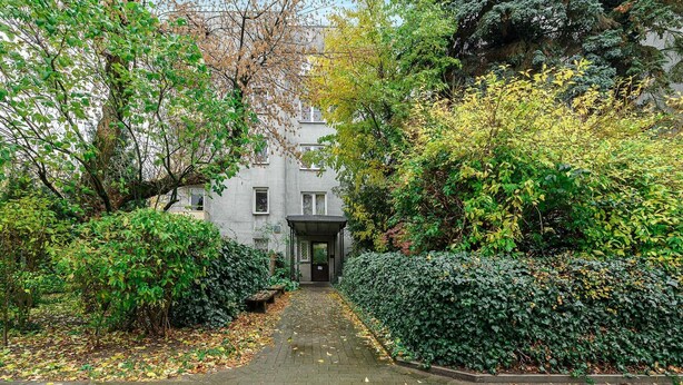 A pathway leading to a building with a covered entrance, surrounded by dense foliage and trees. - Lush Stay on Przasnyska (Warszawa 2)