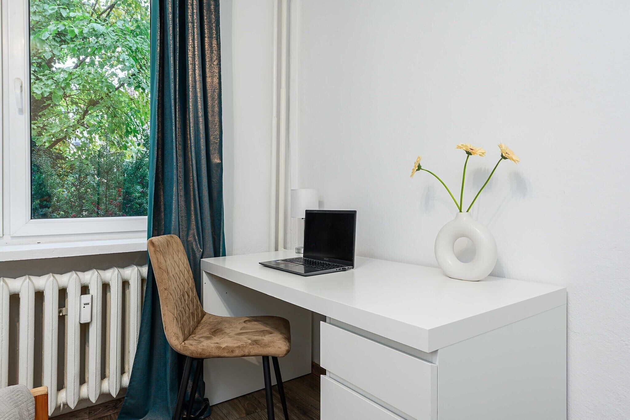 Un espace de bureau moderne avec un bureau blanc, un ordinateur portable, une chaise, un vase avec des fleurs et une fenêtre avec des rideaux.