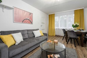 A modern living room with a grey sofa, yellow cushions, a glass coffee table, and a dining area with yellow curtains. - Lush Stay on Przasnyska (Warszawa 2)