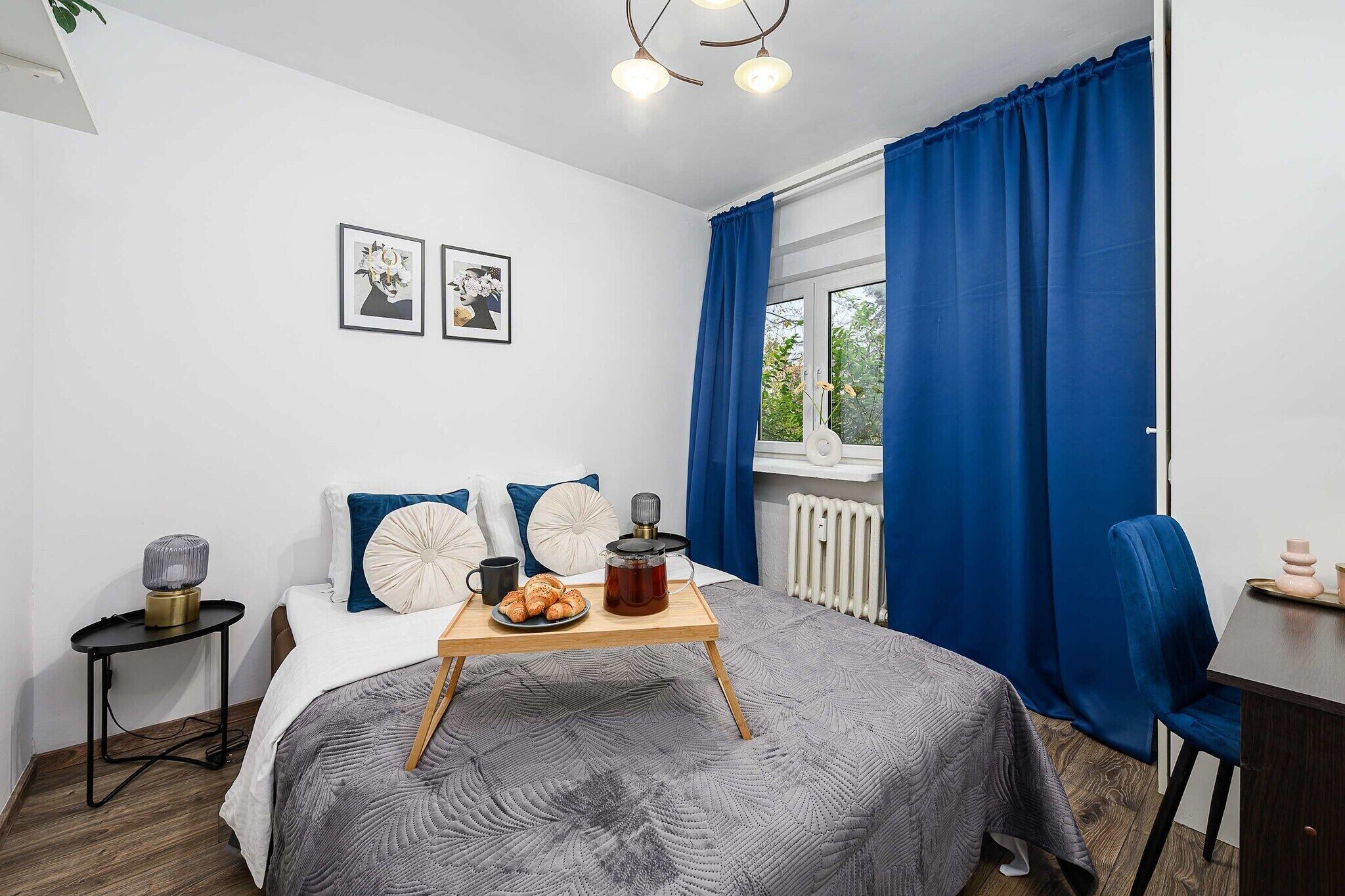 Un dormitorio con cama, mesita de noche con lámpara, ventana con cortinas y una pared con dos cuadros enmarcados.