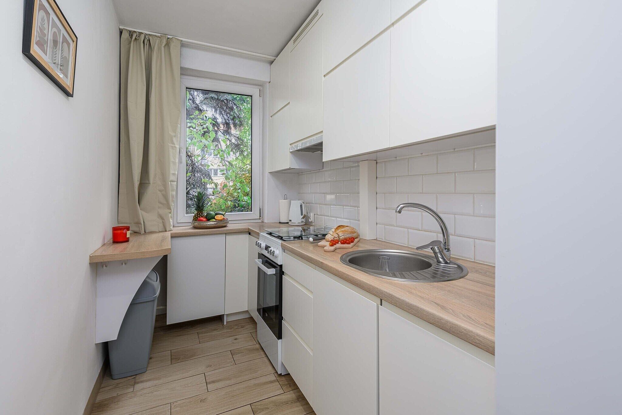 Une cuisine compacte avec des meubles blancs, un plan de travail en bois, un évier en acier inoxydable et une fenêtre donnant sur un espace verdoyant.