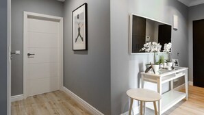Um corredor com uma porta branca, um quadro na parede, uma toucador branca com espelho e um pequeno banquinho.