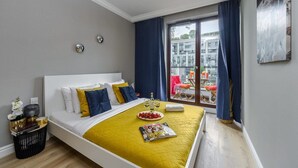 Quarto de hotel com cama, uma mesinha com uma revista, uma garrafa e copos, e uma varanda com mesa e cadeiras.