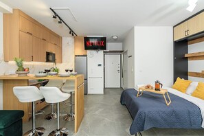 Ein kompaktes Hotelzimmer mit einer Küchenzeile, einem Bett mit blauer Bettdecke, einem Couchtisch und einem an der Wand montierten Fernseher.