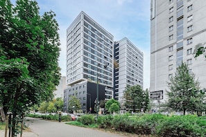 Ein modernes Hochhaus mit markantem Hauseingang an einer Ecke, umgeben von üppiger Bepflanzung und einem gepflasterten Weg.