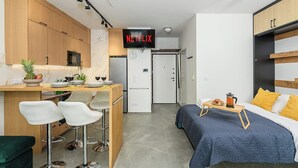 A compact hotel room with a kitchenette, a bed with a blue quilt, a coffee table, and a TV mounted on the wall. - Lush Home near Varso Tower (Warszawa)
