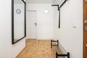 Ein Korridor mit Holzdielenboden, einer weißen Tür und einer Bank mit Metallstreben. An der Wand befinden sich zwei Wanduhren und ein Lichtschalter.