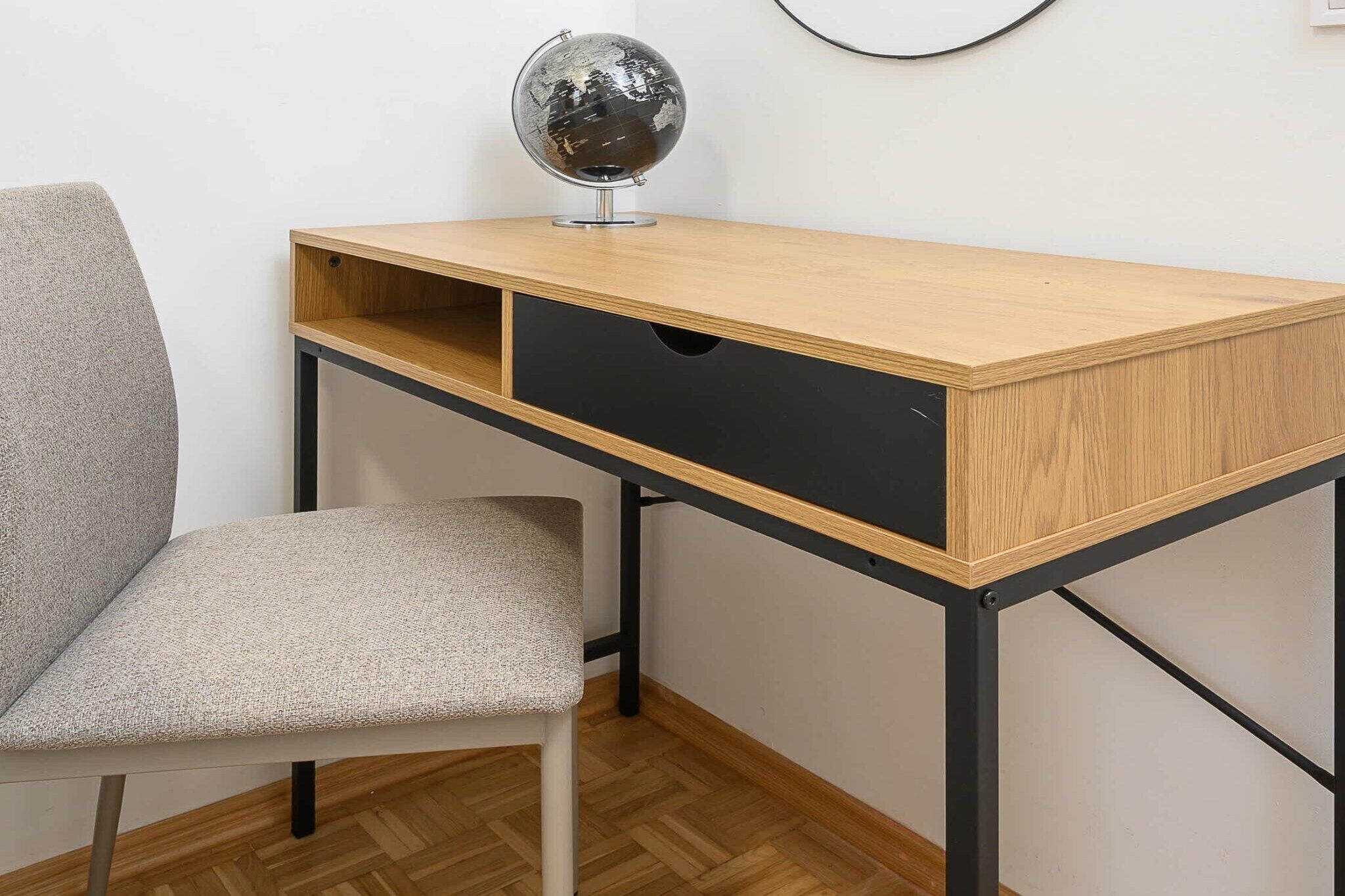 A wooden desk with a black drawer and a globe on top, a grey chair, and a herringbone-patterned floor.