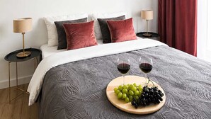 Een netjes opgemaakt bed met een grijs geweven sprei, twee nachtlampjes, en een houten dienblad met druiven en wijnglazen op het bed.