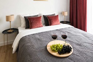 A neatly made bed with a grey textured bedspread, two bedside lamps, and a wooden tray with grapes and wine glasses on the bed. - Renovated Coastal Stone House in Lanildut (Łódź)