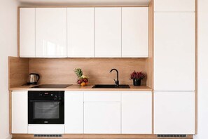 A modern kitchen with white cabinets, a black oven, a sink, and a small countertop with a kettle and decorative items. - Renovated Coastal Stone House in Lanildut (Łódź)