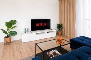 A modern living room with a flat-screen TV displaying 'NETFLIX', a white entertainment unit, a glass coffee table with magazines and croissants, blue sofas, and potted plants. - Renovated Coastal Stone House in Lanildut (Łódź)