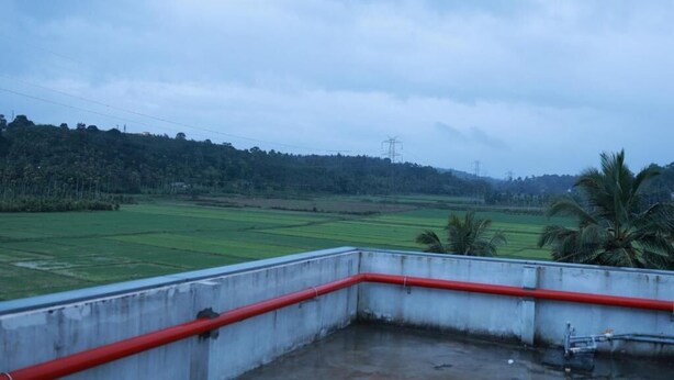 A view from a balcony with a concrete floor and red railing, overlooking a green field with palm trees and a distant power line. - MASBAH RESIDENCY (Mananthavady)