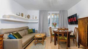 A living room with a sofa, a dining table with chairs, a TV mounted on the wall, and a window with curtains. - Beachfront Villa with Panoramic Sea Views (Warszawa 2)