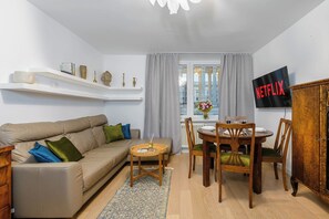 Ein Wohnzimmer mit einer Couch, einem Esstisch mit Stühlen, einem an der Wand befestigten Fernseher und einem Fenster mit Vorhängen.