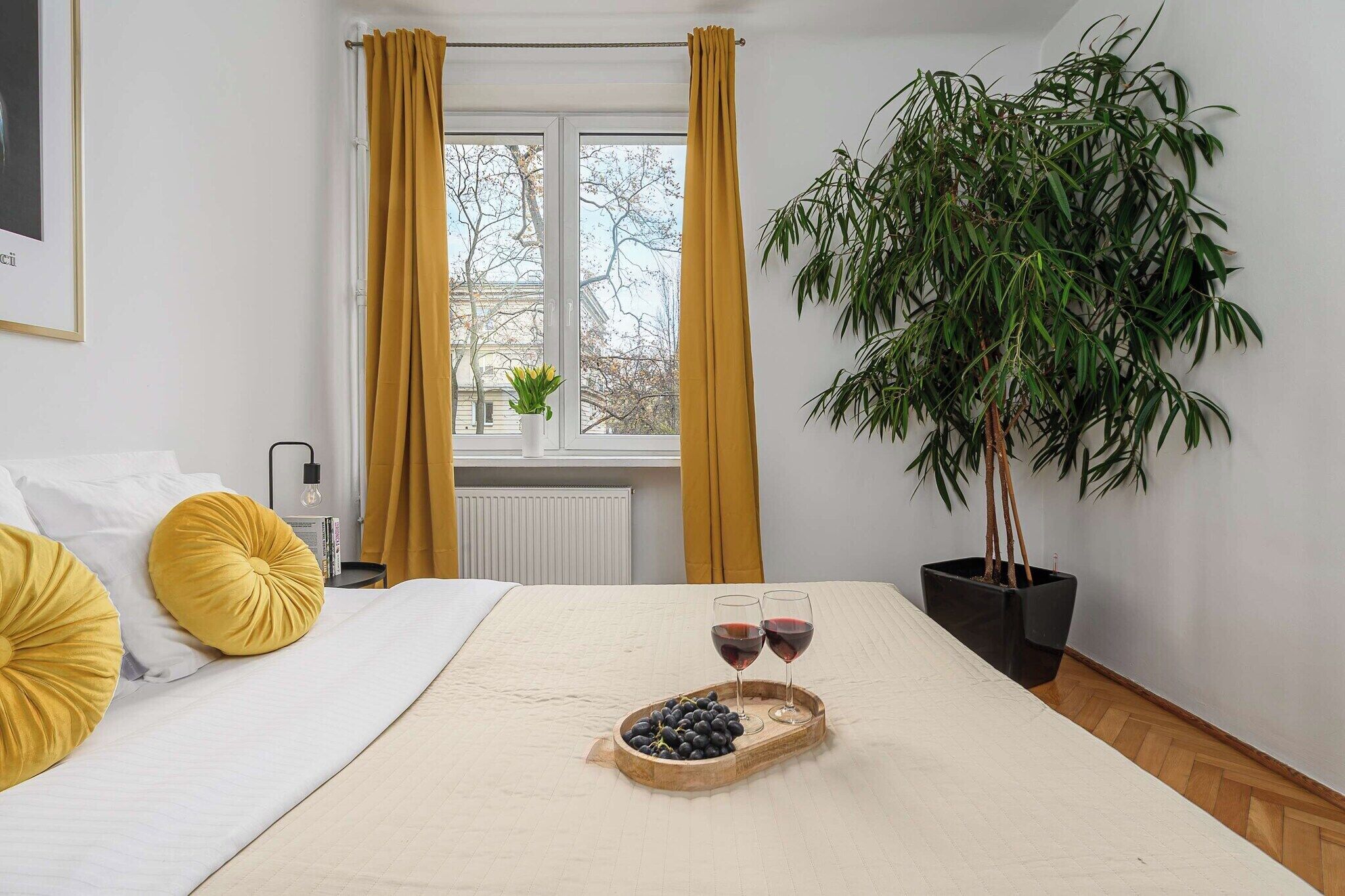 Un dormitorio con cama, almohadas amarillas, mesita de noche con lámpara, ventana con cortinas, planta en maceta y bandeja con uvas y copas de vino.