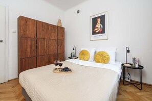 Ein Schlafzimmer mit einem Bett, einem Holzschrank, einem gerahmten Kunstwerk, Nachttischen mit Lampen sowie einem Tablett mit Weingläsern und Trauben.