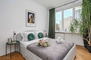 A neatly arranged bedroom with a bed, pillows, a bedside table, a window with curtains, a potted plant, and a framed picture on the wall. - Warsaw CIty Apartment with Paid Parking (Warszawa)