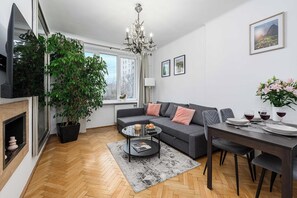 Un salon moderne avec un canapé gris, une table basse, une table à manger pour deux personnes, une grande plante en pot et un lustre.