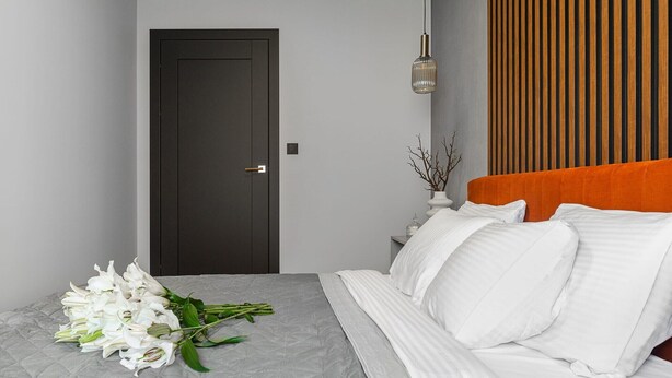 Hotelkamer met een bed, een deur, een nachtkastje, een vaas met takken en een wand met verticale houten lamellen.