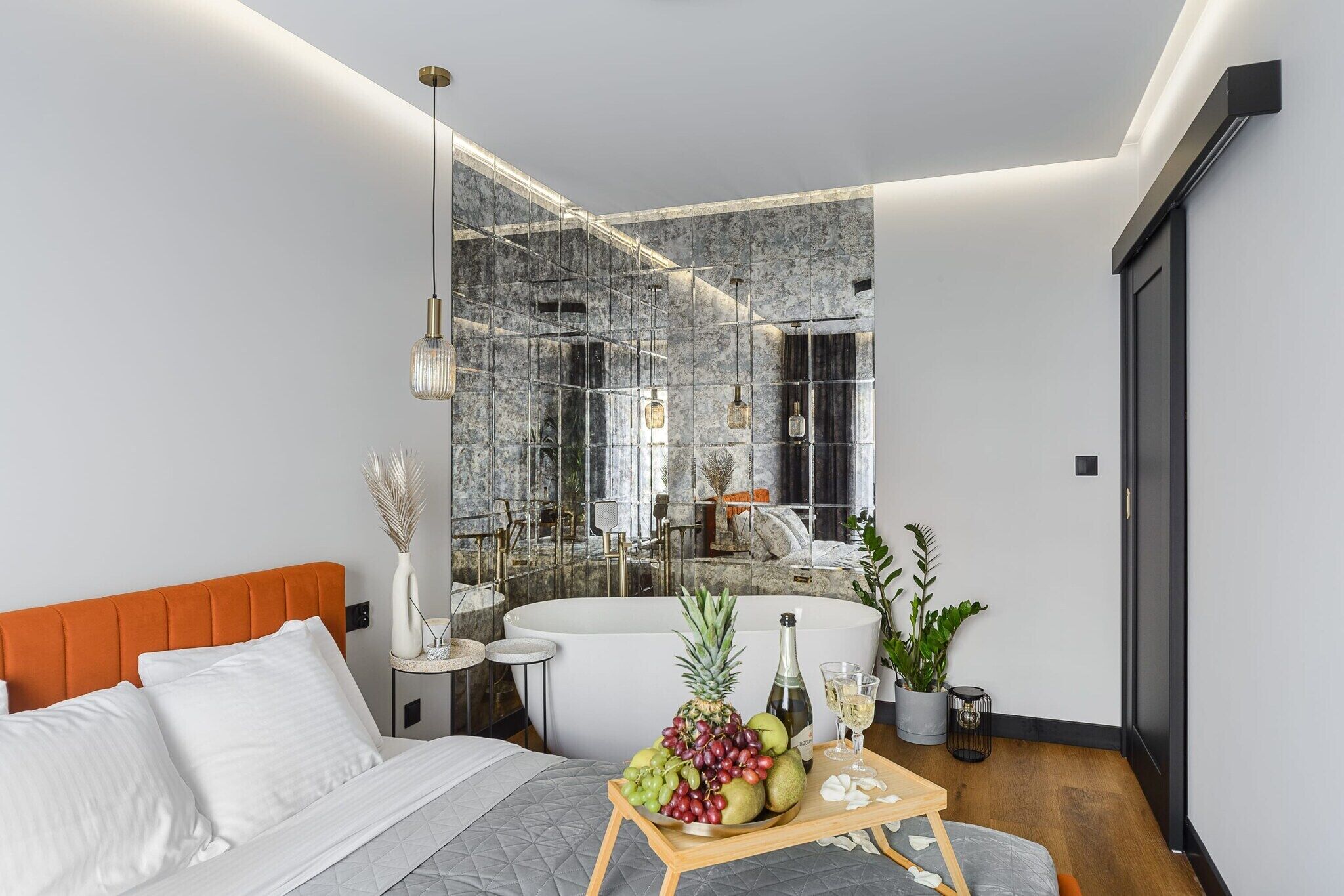 Une chambre à coucher moderne avec un lit, une table de chevet sur laquelle se trouve un panier de fruits, un miroir et une salle de bain avec une baignoire.